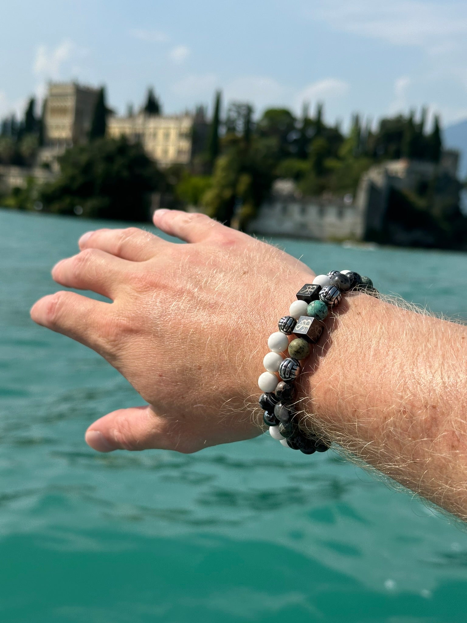 Twee kralenarmbanden met donkere en veelkleurige stenen liggen op een grijs oppervlak, naast een stuk drijfhout. Elke armband heeft een klein vierkant bedeltje met initialen.