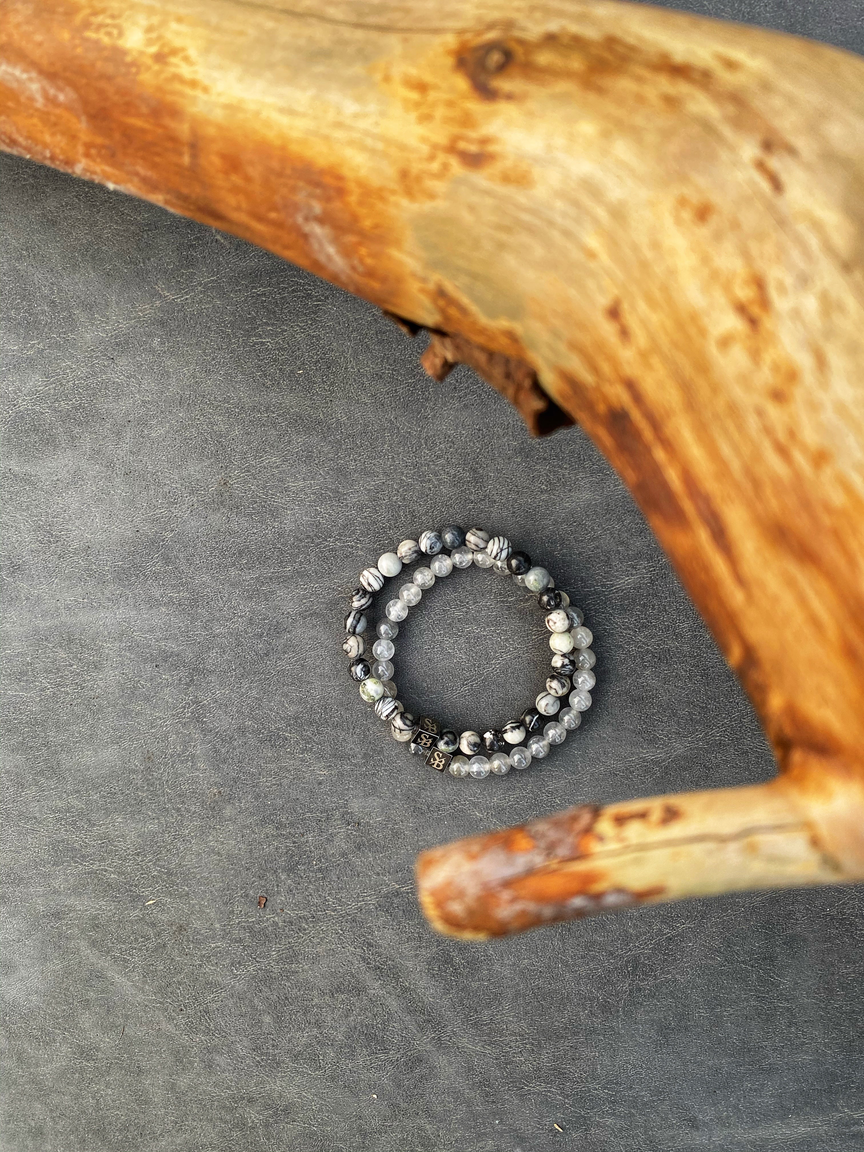 Twee kralenarmbanden met grijze en witte gemarmerde stenen zijn op een donkergrijs oppervlak geplaatst. Een stuk gebogen, getextureerd hout bevindt zich in het bovenste deel van de afbeelding.