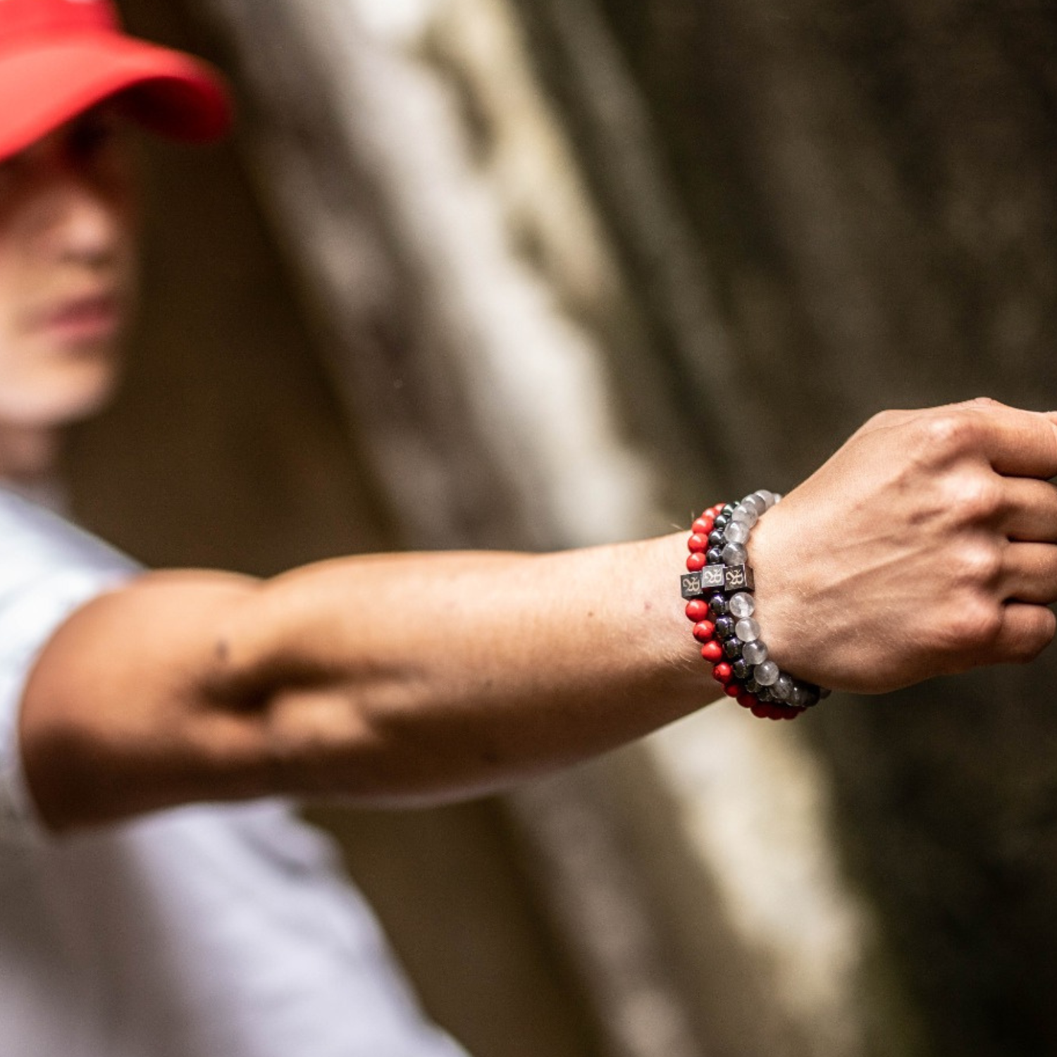 Een persoon met een rode pet wijst met zijn hand, en toont armbanden van natuursteen en edelsteen in rode, grijze en zwarte tinten om zijn pols. De achtergrond is wazig, waardoor de arm en de armbanden worden benadrukt. De persoon draagt een wit shirt.