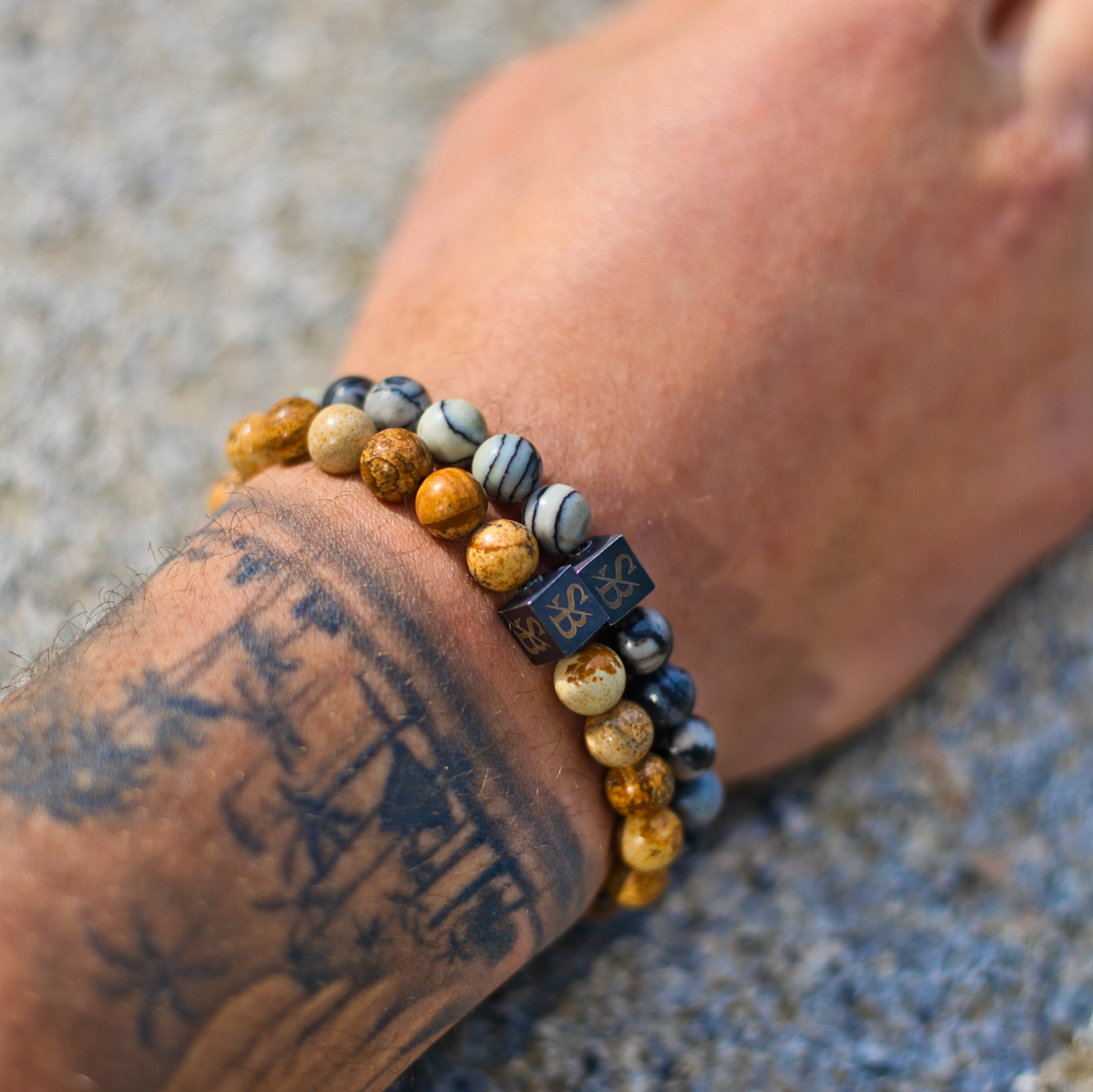 Een getatoeëerde onderarm draagt twee kralenarmbanden, waaronder de Stoney Bracelets Desert Jasper Stones (8mm), tegen een gestructureerde, lichtgekleurde achtergrond.