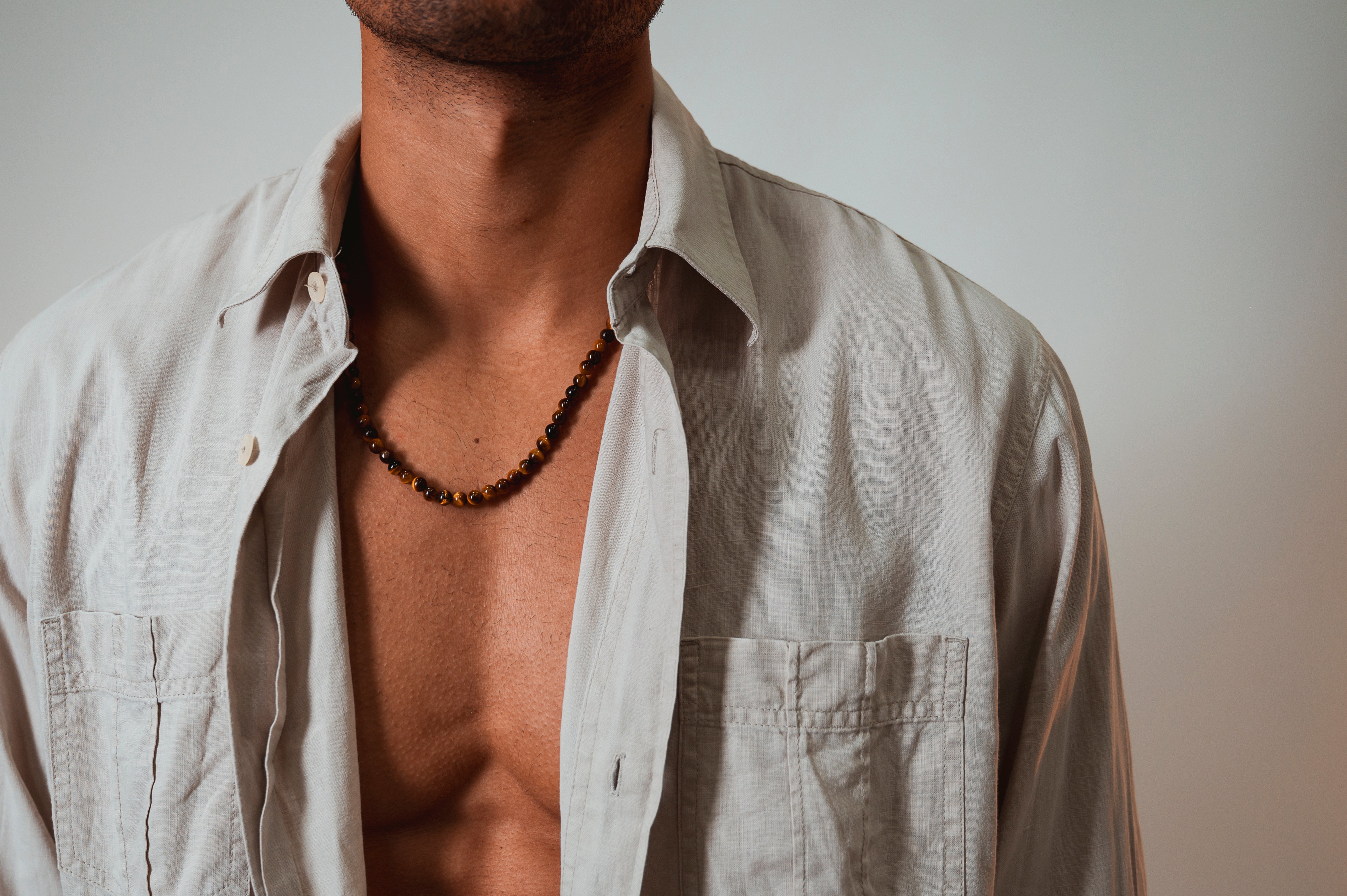 Drie kralenkettingen worden op een stenen oppervlak tentoongesteld. De kettingen hebben grijze, bruine en blauwe kralen, elk met een gouden sluiting en een klein rond label.