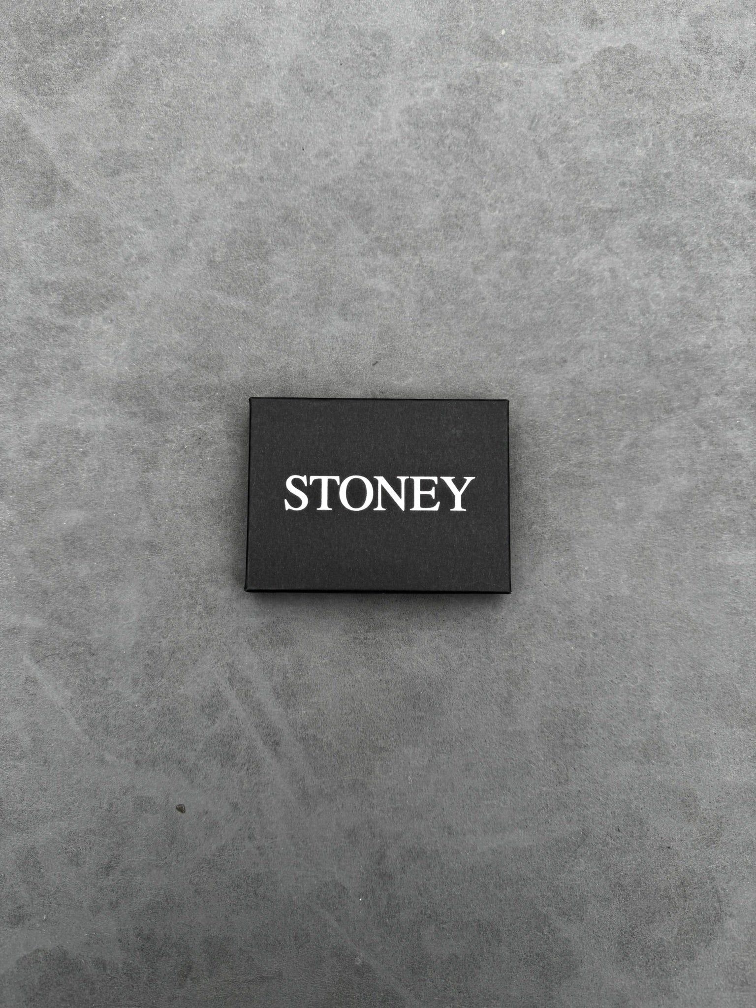Een platte, rechthoekige, zwarte kaart met het woord "STONEY" in witte hoofdletters, geplaatst op een grijs oppervlak.
