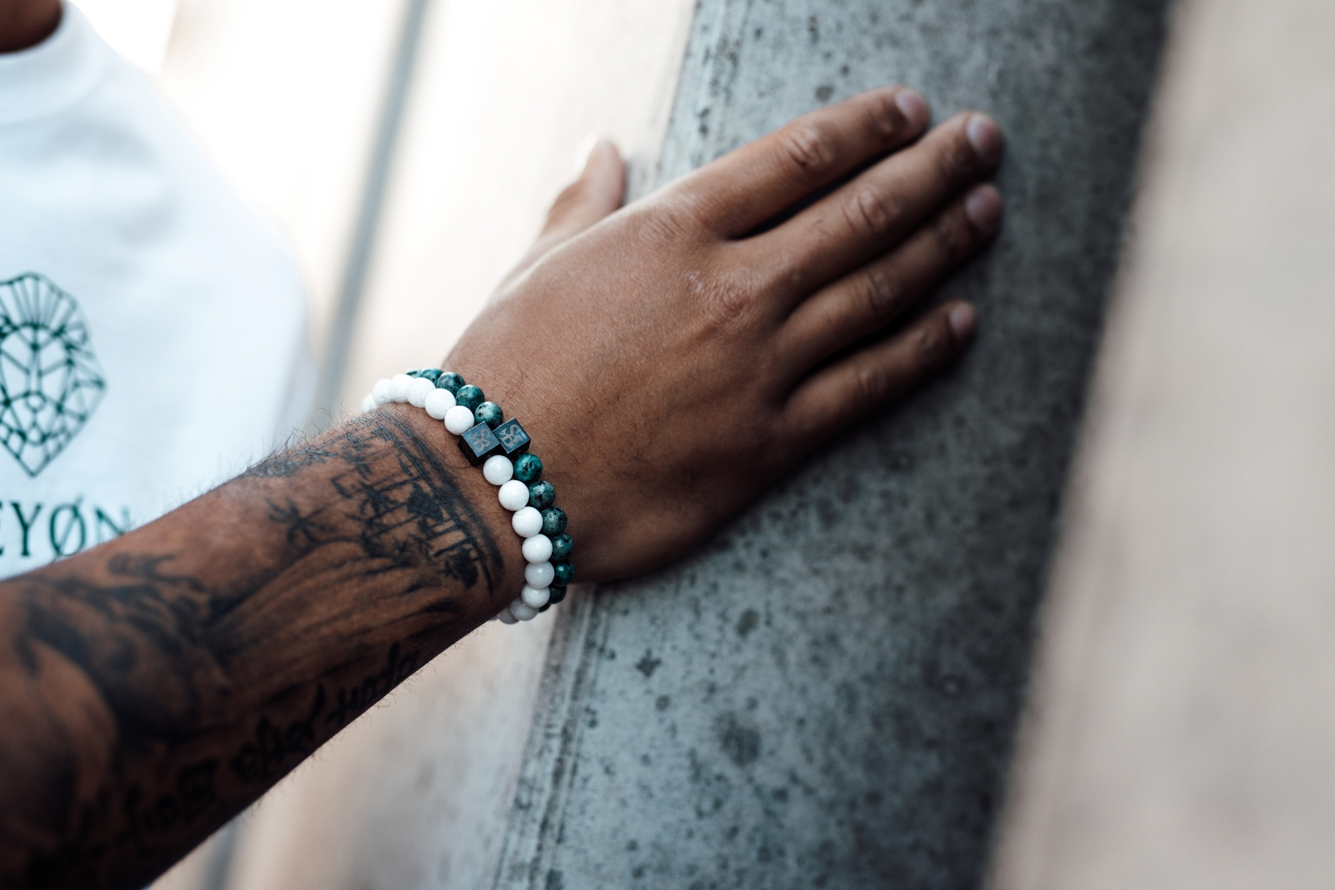 Een getatoeëerde arm rust op een betonnen oppervlak. De persoon draagt twee kralenarmbanden, een witte en een groene en zwarte. Een gedeeltelijke weergave van een wit shirt met een logo is zichtbaar.