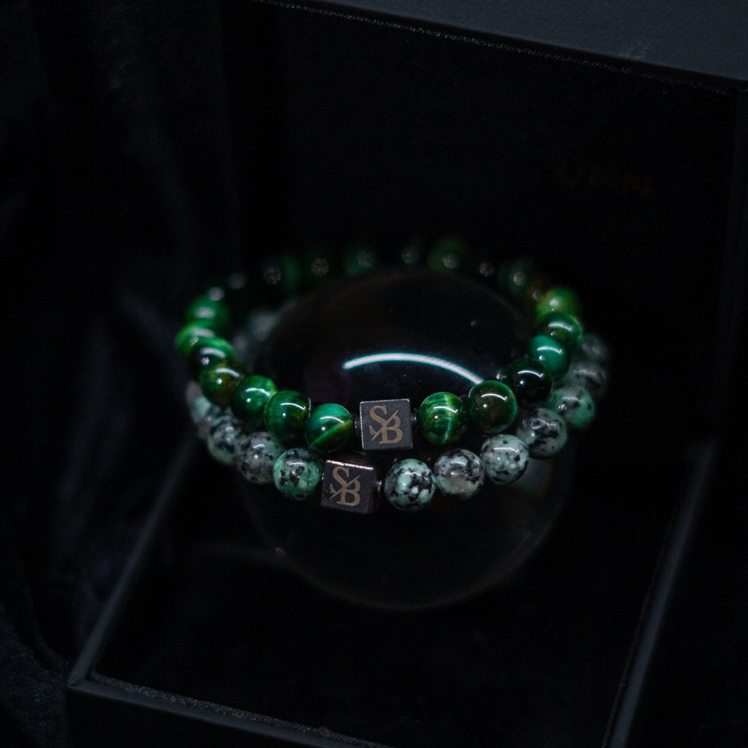 Een close-up van twee kralenarmbanden van Stoney Bracelets. De ene is versierd met groene tijgeroogstenen (8 mm), terwijl de andere is versierd met zwarte en witte gespikkelde stenen. Elke armband heeft een klein vierkant bedeltje met de initialen "SB" gegraveerd en wordt weergegeven op een strak zwart oppervlak.