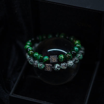 Een close-up van twee kralenarmbanden van Stoney Bracelets. De ene is versierd met groene tijgeroogstenen (8 mm), terwijl de andere is versierd met zwarte en witte gespikkelde stenen. Elke armband heeft een klein vierkant bedeltje met de initialen "SB" gegraveerd en wordt weergegeven op een strak zwart oppervlak.