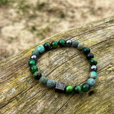 Een armband van Stoney Bracelets, genaamd "Swamp | Mixed Stones (8mm)", met groene, blauwe en zwarte ronde kralen, wordt op een houten oppervlak getoond. Deze elegante armband bevat een opvallende kleine zwarte vierkante kraal met een geëtst ontwerp. De achtergrond is wazig, wat een aardse buitenomgeving laat zien.