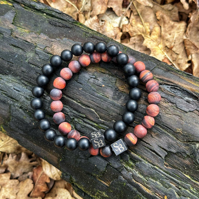 Twee kralenarmbanden van Stoney Bracelets met vierkante metalen bedels gegraveerd met "S&B" rusten op een met mos bedekte boomstam omringd door gevallen bladeren. Eén armband heeft gladde zwarte kralen, terwijl de andere afwisselt tussen Matte Obsidian Stones (8mm) en roodbruine natuurstenen kralen met strepen.