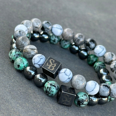 Twee Stoney Bracelets "Nautic | Mixed Stones (8mm)" armbanden worden op een grijs oppervlak getoond. Elk heeft een mix van gemarmerde witte, grijze, groene en zwarte natuursteen kralen. Beide hebben een kubusvormige kraal met de letters "SB" erin gegraveerd. De kralen variëren in textuur en patroon.
