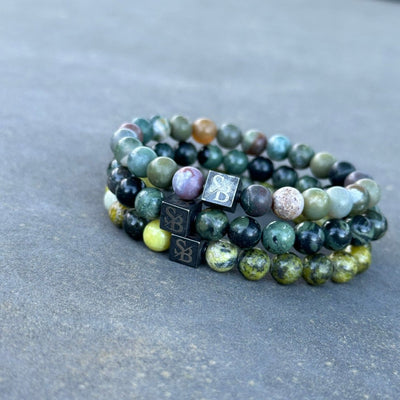 Drie armbanden uit de Stoney Bracelets' Triple Green Set, gemaakt met 8mm kralen in verschillende tinten groene, blauwe en bruine edelstenen, zijn gestapeld op een grijs oppervlak. Elke armband heeft een klein zilveren vierkant bedeltje gegraveerd met de initialen "SB.