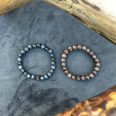 Twee elegante kralenarmbanden van Stoney Bracelets' Premium Coffee Set (8mm) rusten op een grijs oppervlak, delicaat omzoomd door houten takken. De armband aan de linkerkant toont kleine, donkergrijze kralen met een natuurlijke stenen uitstraling, terwijl de armband aan de rechterkant grotere kralen met een opvallend bruin en wit gemarmerd patroon heeft. Beide stukken zijn versierd met een kleine vierkante bedel gegraveerd met de letters "SB.