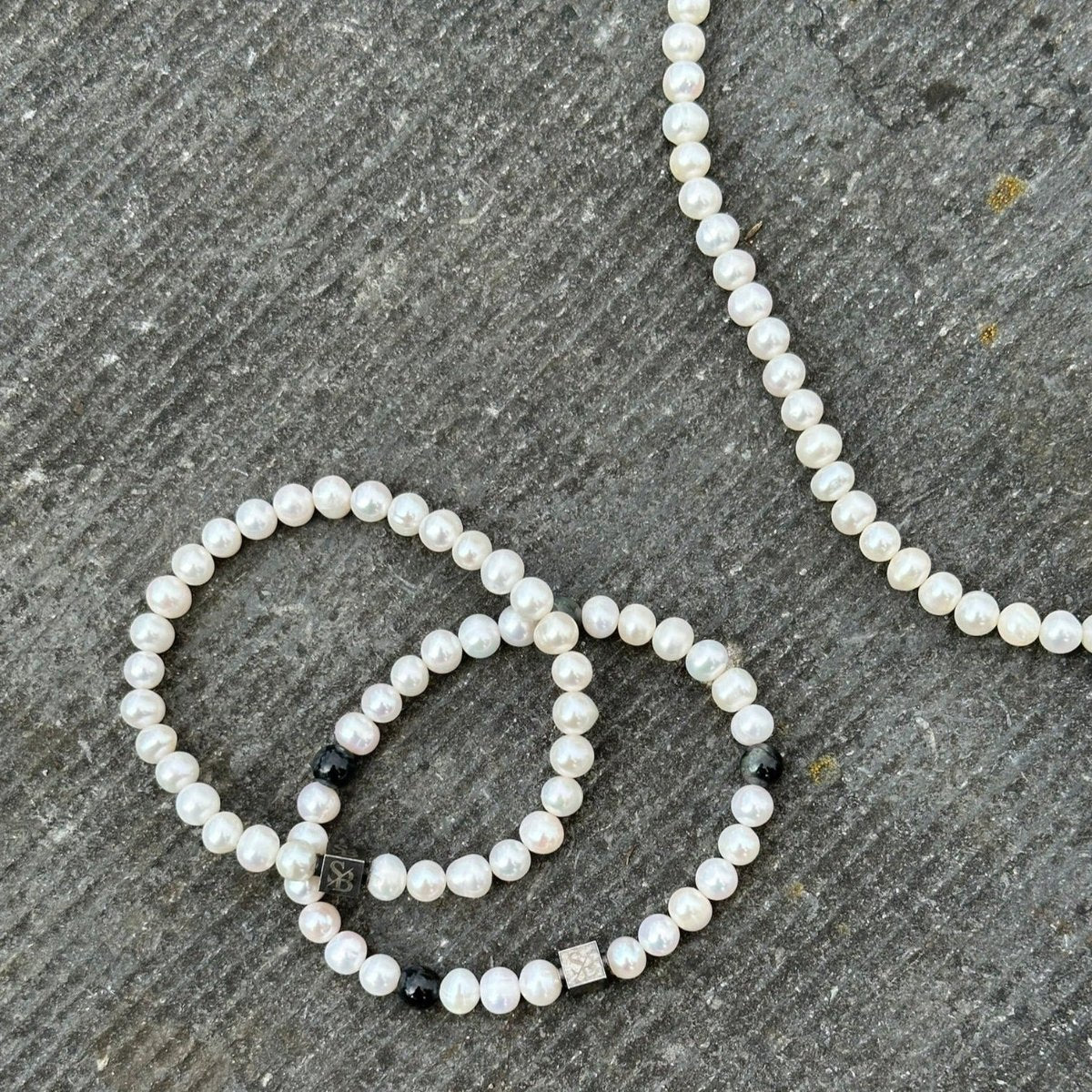 Close-up van een parelsieraad op een getextureerd, grijs oppervlak. De set bestaat uit een parelketting en twee Stoney Bracelets' Rounded Pearl Bracelet I (7mm). De armbanden zijn versierd met kleine zwarte en zilveren decoratieve kralen die tussen de parels zijn verspreid, wat een vleugje elegantie toevoegt met kralen en natuursteenelementen.