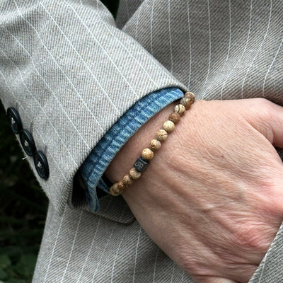 Een persoon met een bruin krijtstreepjasje en een blauw denim overhemd draagt een armband van Stoney Bracelets Matte Desert Jaspis Stones (6 mm). De bruine en beige tinten van de unisex natuurstenen armband passen bij de drie zwarte knopen van het jasje.