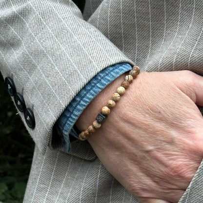 Een persoon met een bruin krijtstreepjasje en een blauw denim overhemd draagt een armband van Stoney Bracelets Matte Desert Jaspis Stones (6 mm). De bruine en beige tinten van de unisex natuurstenen armband passen bij de drie zwarte knopen van het jasje.