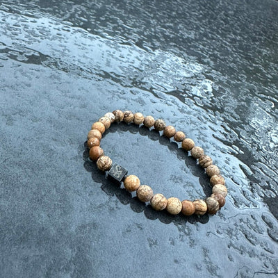 Een Stoney Bracelets Matte Desert Jaspis Stones (6mm) armband met bruin gemarmerde kralen en een klein zwart rechthoekig bedeltje rust op een nat, gestructureerd grijs oppervlak.