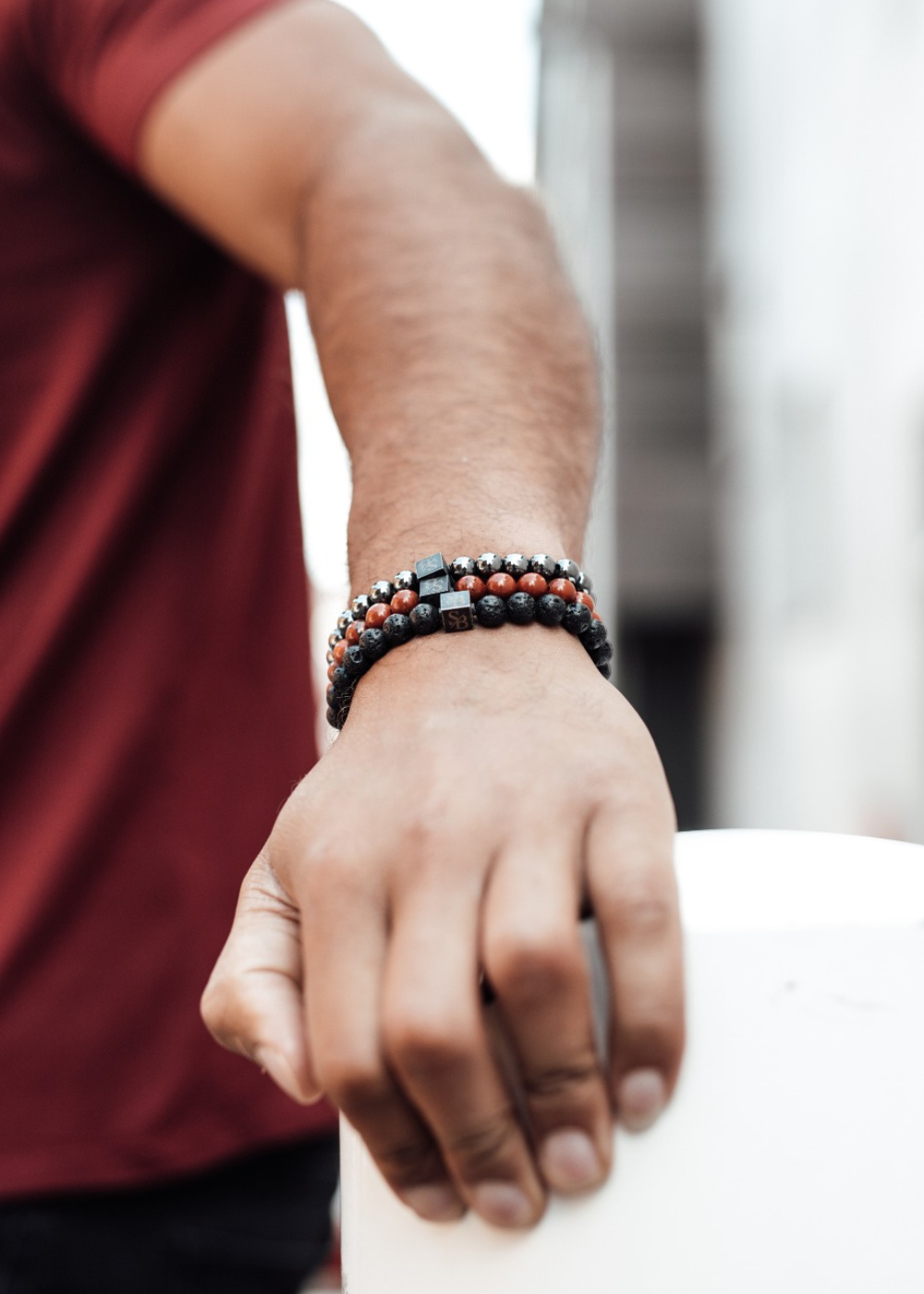 Een persoon die een rood shirt draagt, toont zijn linkerarm versierd met een armband van meerdere kralenarmbanden in zwart, rood en zilver. Zijn hand rust op een wit oppervlak. De achtergrond is wazig, waardoor de aandacht op de arm en de armbanden wordt gevestigd.