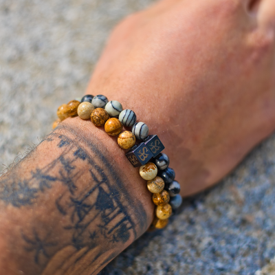 Een getatoeëerde onderarm draagt twee kralenarmbanden, waaronder de Stoney Bracelets Desert Jasper Stones (8mm), tegen een gestructureerde, lichtgekleurde achtergrond.