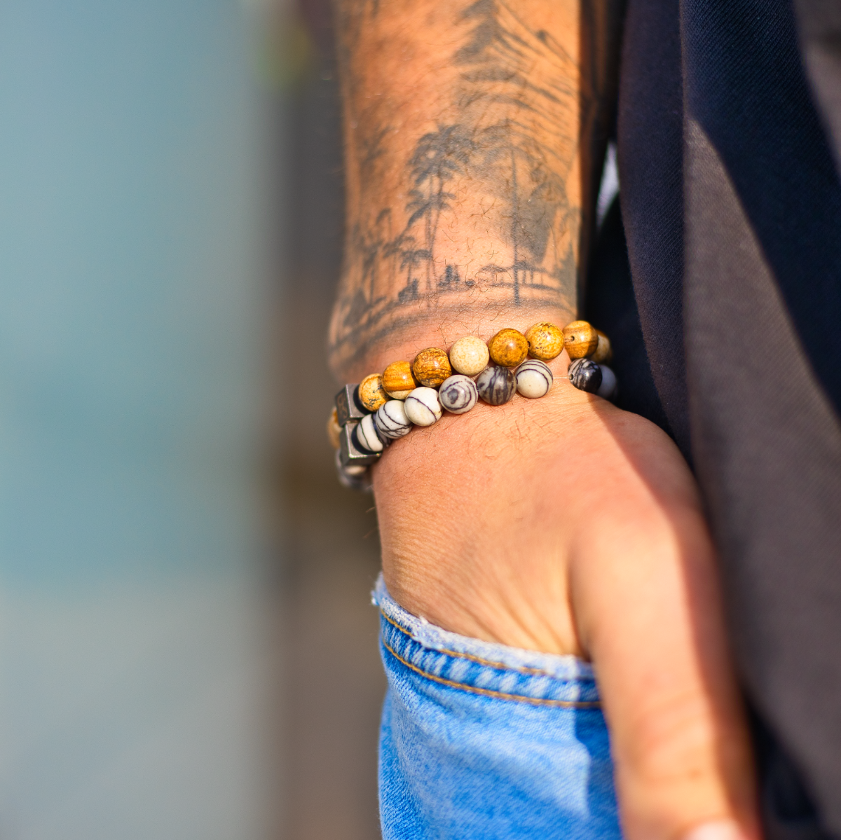Iemand met een tatoeage van een boom op zijn onderarm draagt een Stoney Bracelets Desert Jasper Stones (8mm) armband van bruin en wit gemarmerde kralen. Hij draagt zijn hand in een lichtblauwe spijkerbroek en combineert deze met een donker overhemd.