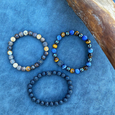 Drie armbanden van Stoney Bracelets "Desert | Mixed Stones (8mm)" staan in een driehoek op een blauw gestructureerd oppervlak naast hout. Elk elastisch, unisex ontwerp biedt een veelzijdige stijl voor dagelijks gebruik.