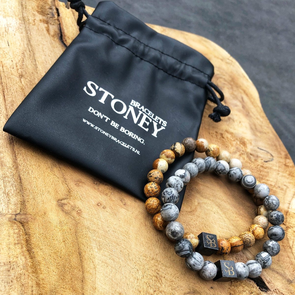 Twee kralenarmbanden gemaakt met 8 mm woestijn Jaspis stenen in aardebruin, grijs en beige liggen op een houten oppervlak naast een zwart zakje met de tekst "Stoney Bracelets. Wees niet saai.