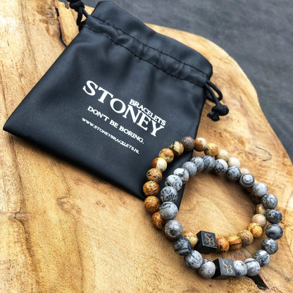 Twee kralenarmbanden gemaakt met 8 mm woestijn Jaspis stenen in aardebruin, grijs en beige liggen op een houten oppervlak naast een zwart zakje met de tekst "Stoney Bracelets. Wees niet saai.